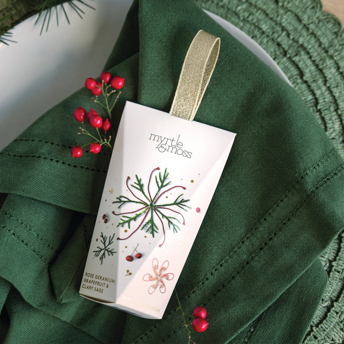 Myrtle & Moss Rose Geranium hanging hand cream box with a gold ribbon loop, printed with a red and green snowflake and red berries, on a green napkin.