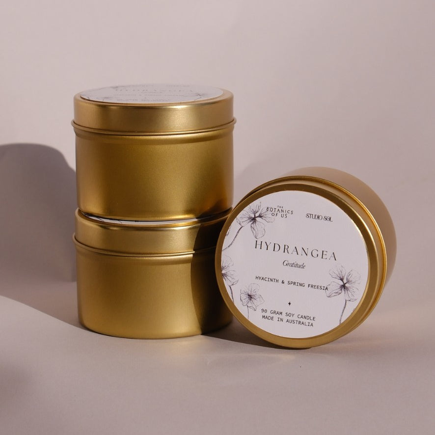 Three gold Hydrangea travel tins, one tipped forward to show its lid label with a drawing of the hydrangea.
