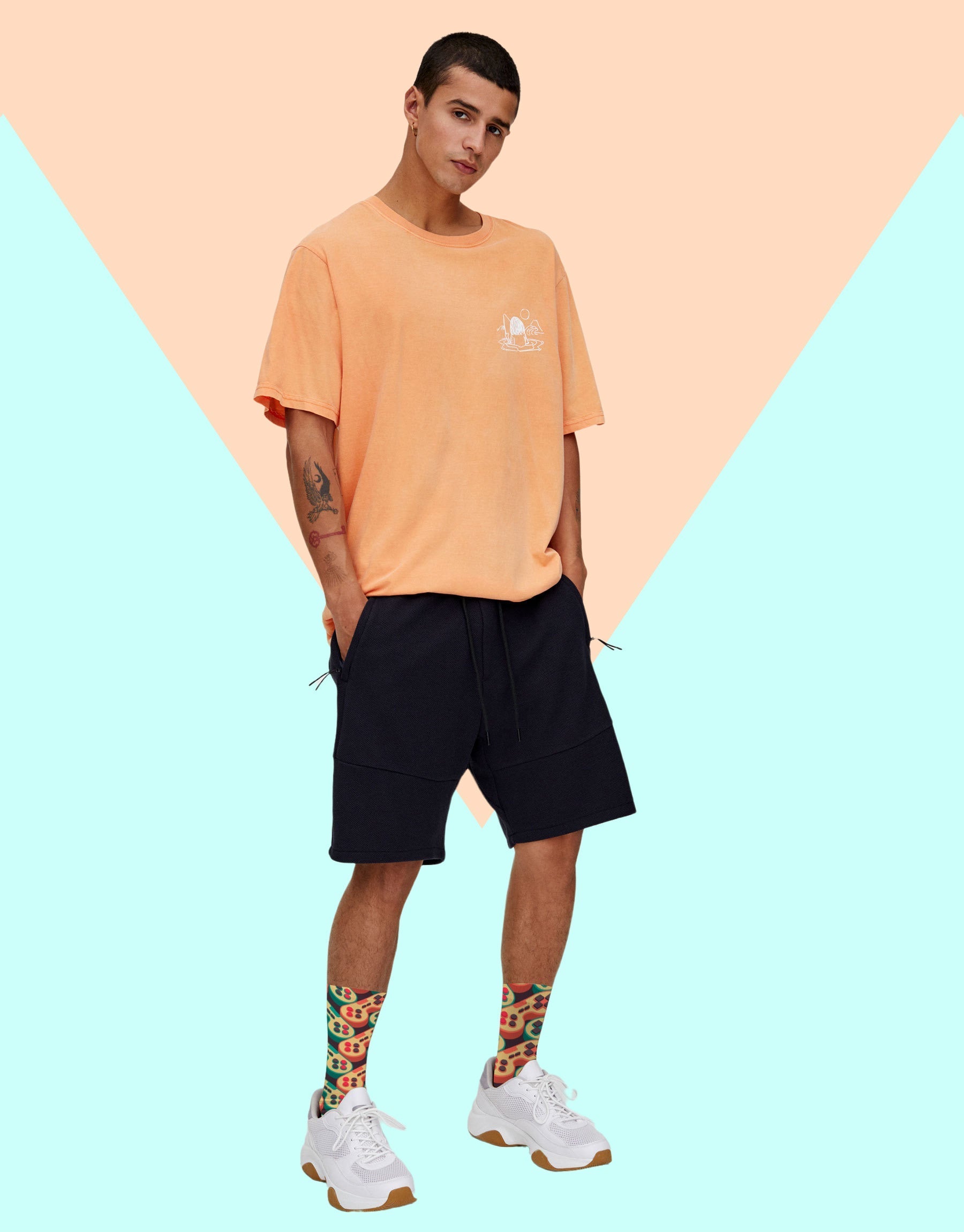 Man in a peach t-shirt and black shorts standing against a peach and pale blue background, wearing white sneakers and the game controller socks.