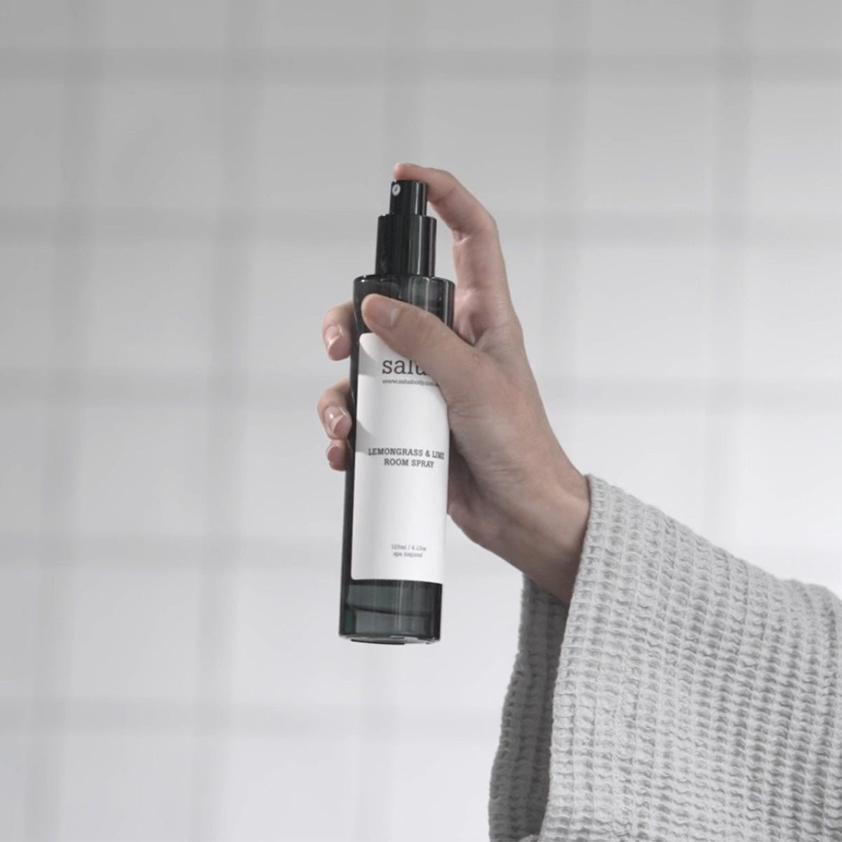 A hand spraying the smoked glass Lemongrass & Lime Room Spray in front of a tiled wall.