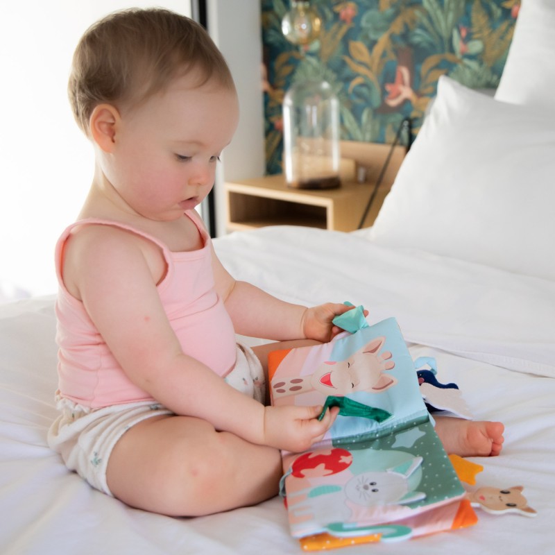 Baby in a pink top sitting on a bed and turning the pages of the soft book.