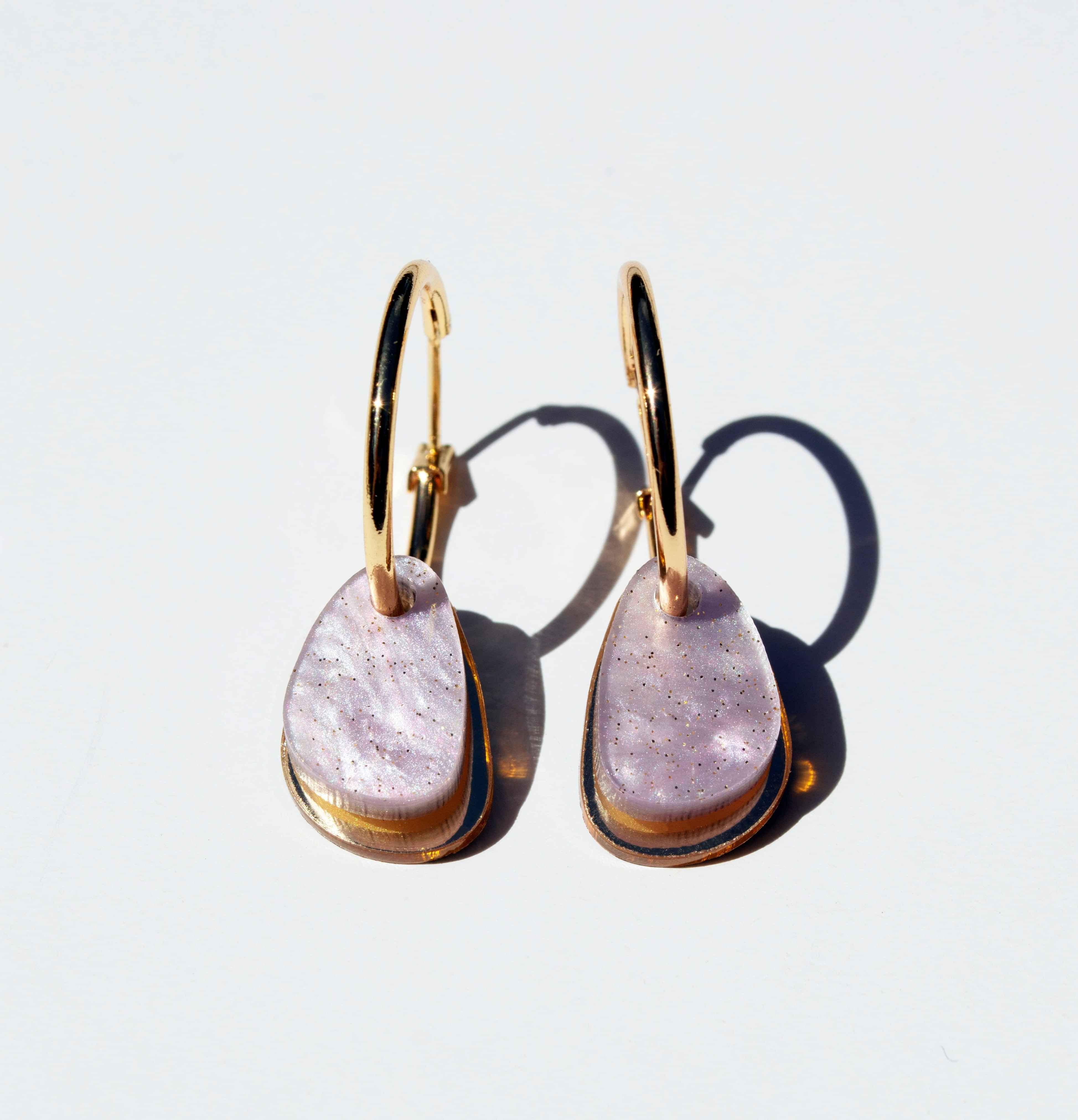 A pair of gold hoop earrings, each hung with a smooth organic teardrop of soft lilac marbled acrylic over a slim gold mirror panel.