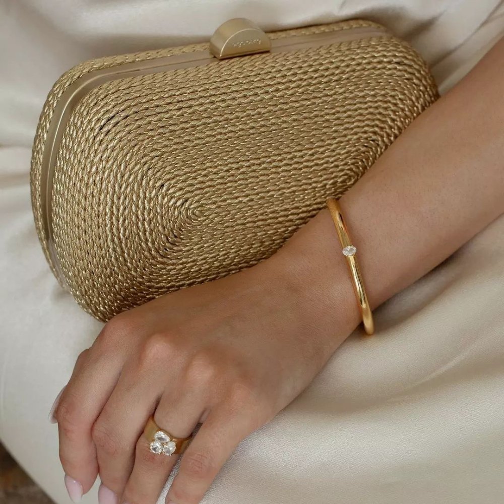 Hand wearing a gold bangle and ring resting on the gold clutch against cream satin