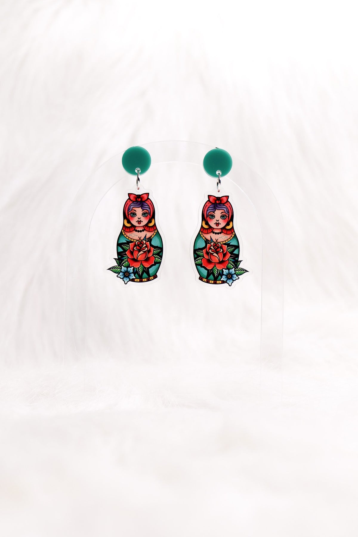 The Matryoshka earrings hanging on a clear stand: green studs above painted nesting dolls.