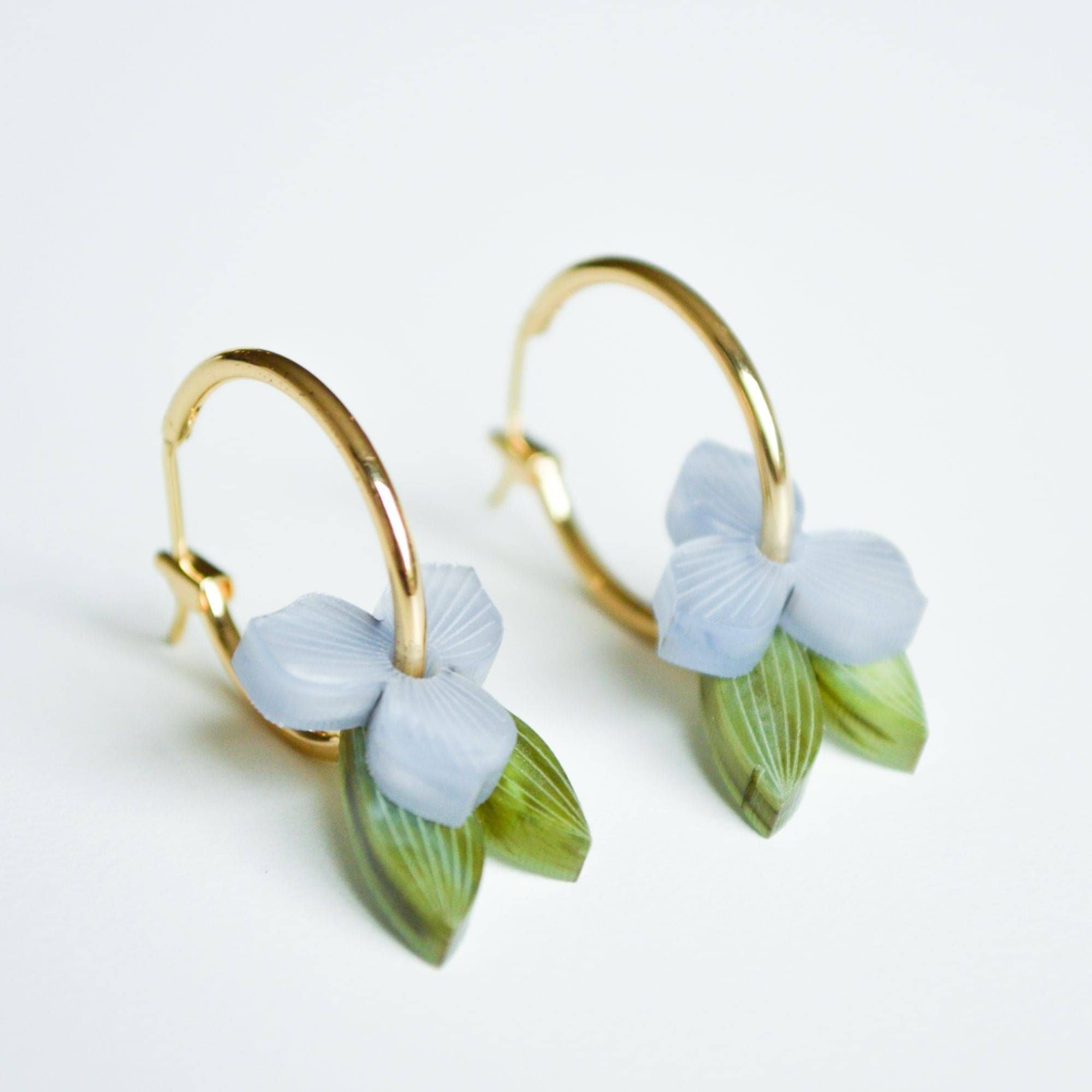A pair of gold hoop earrings, each hung with a small pale periwinkle blue flower and a slender green leaf in marbled acrylic.