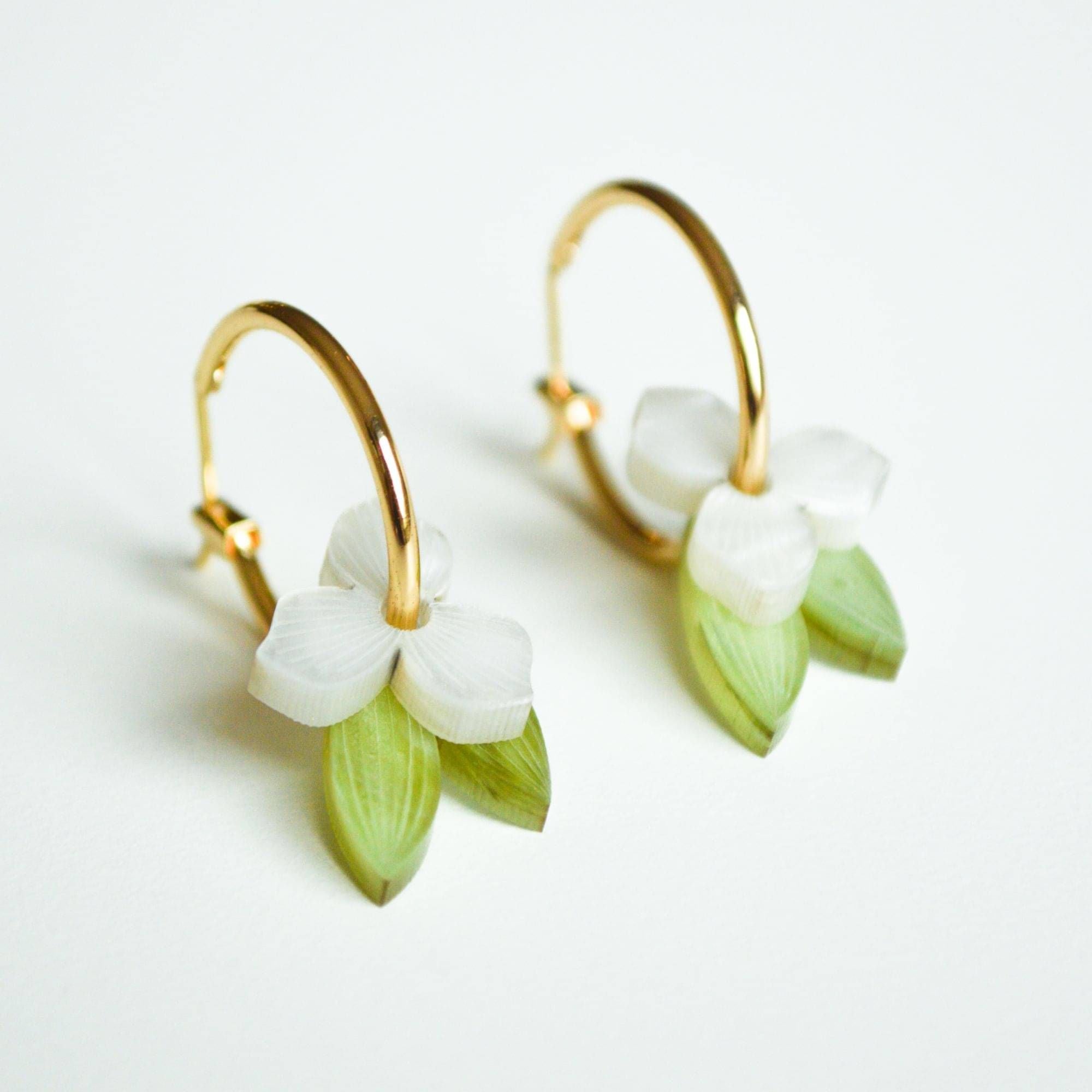 A pair of gold hoop earrings, each hung with a small white flower and a slender green leaf in marbled acrylic.