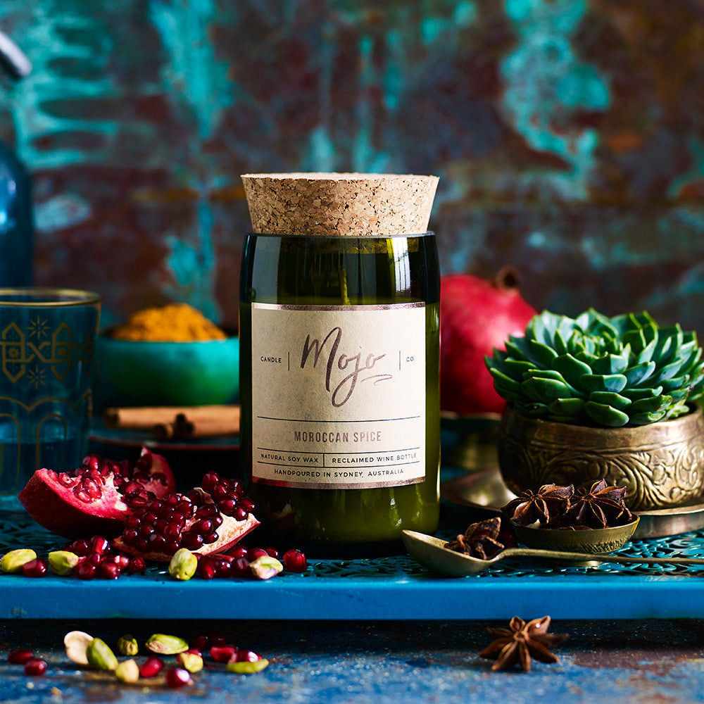 Moroccan Spice candle on a teal setting with pomegranate seeds, a succulent, a brass pot and pistachios