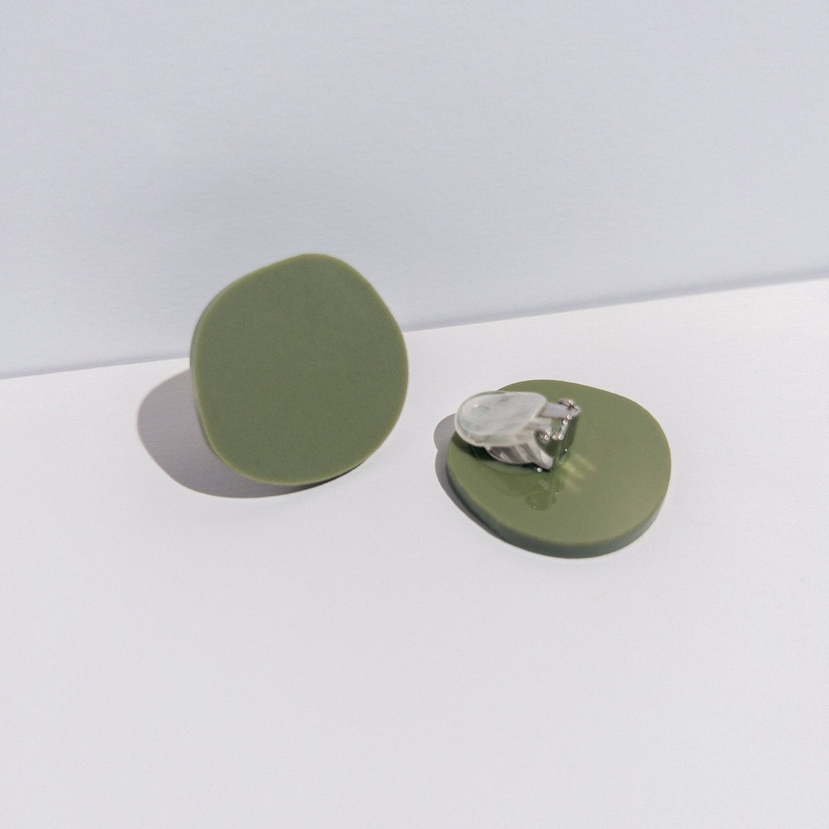 A pair of olive green acrylic disc clip-on earrings, one turned over to show the clip.