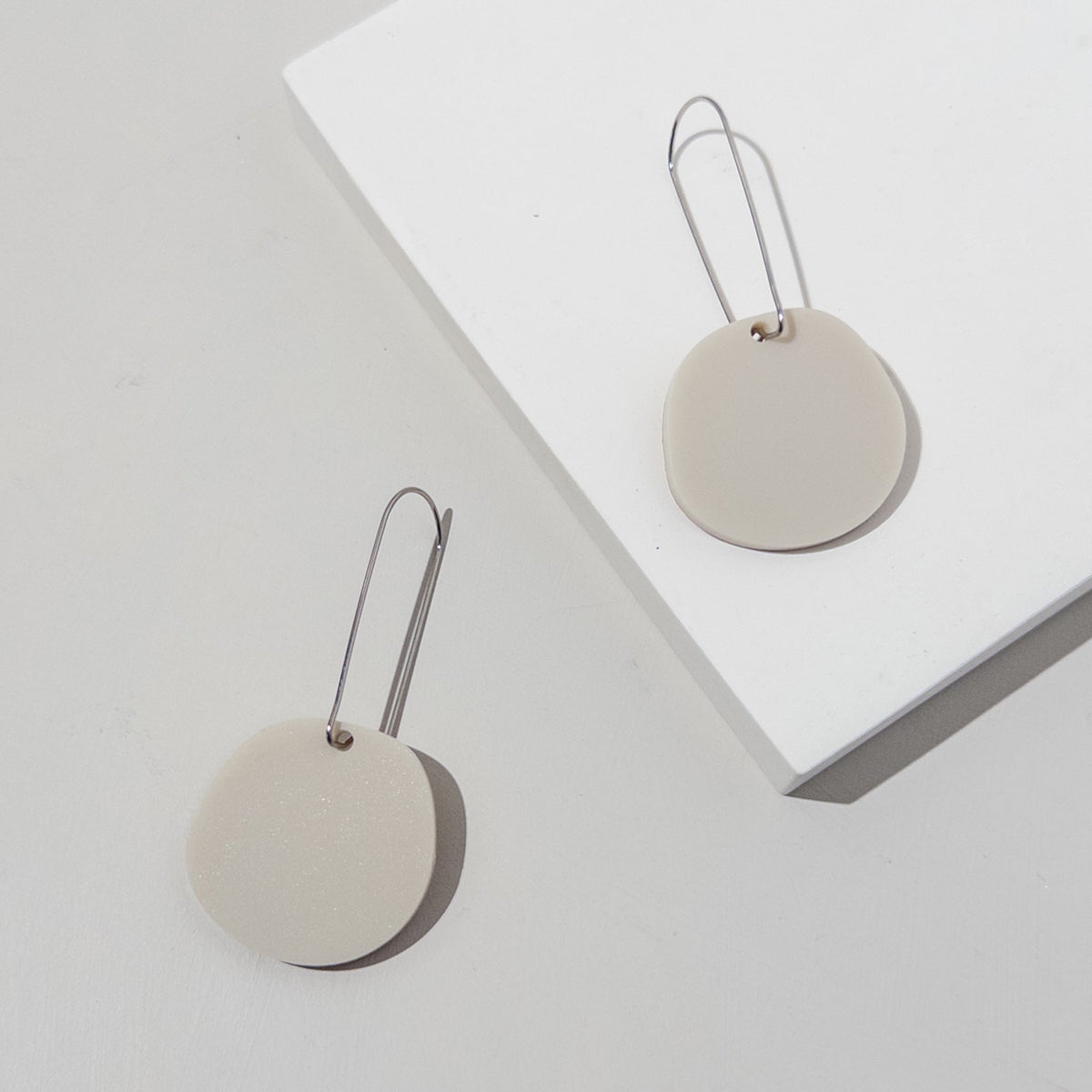 Two ivory beige acrylic discs on long, slim silver-tone ear wires, laid across a white block.