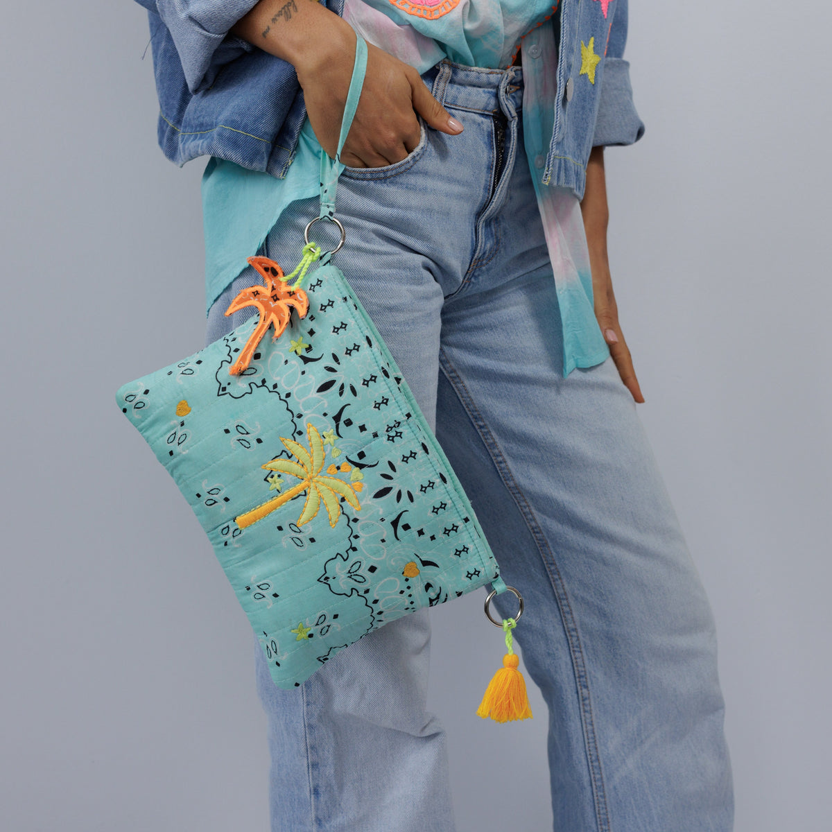 The aqua palm tree pouch carried by its wrist strap against light denim jeans.