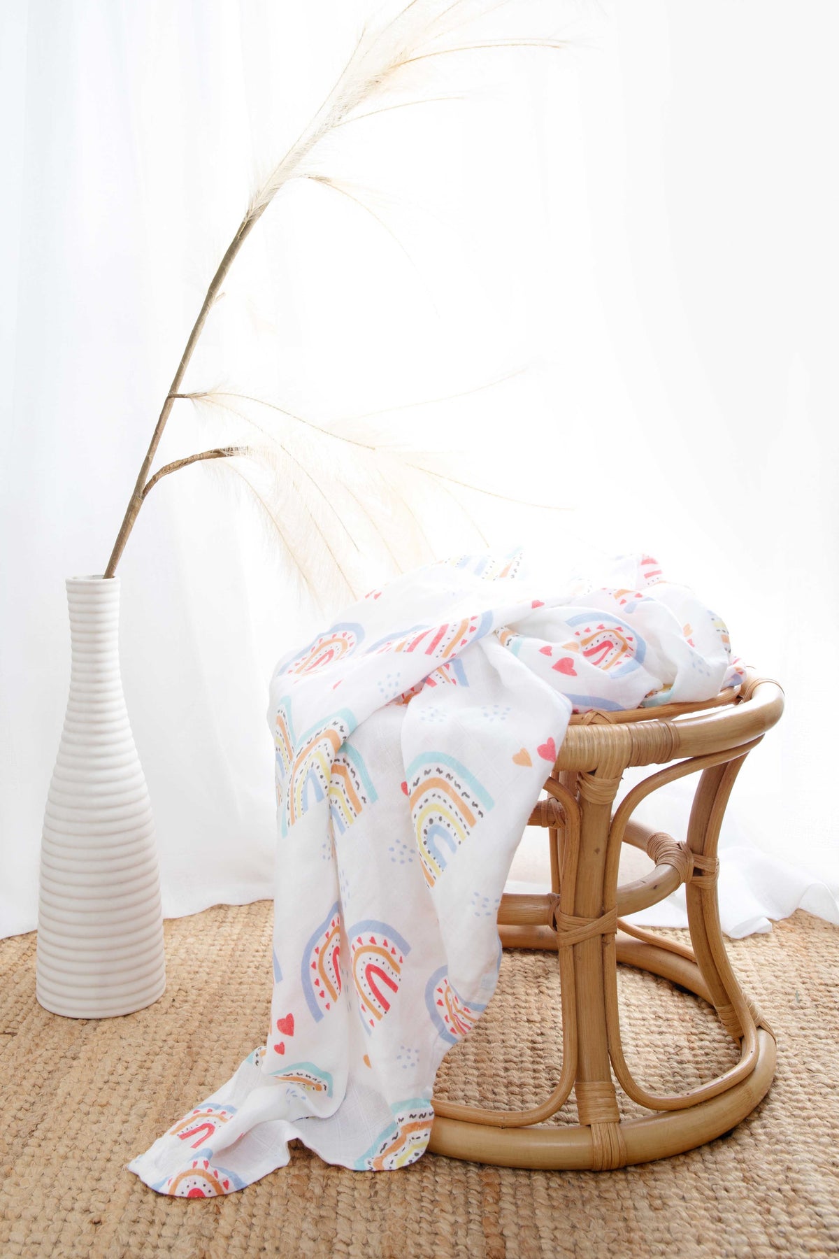 The rainbow-print muslin wrap draped over a rattan stool beside a white vase of pampas grass.