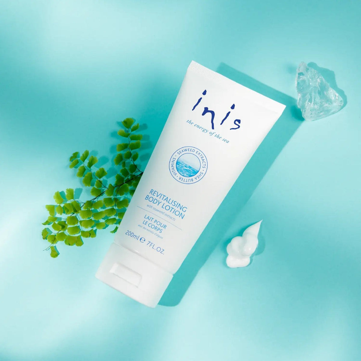 The white Inis 200 ml lotion tube lying on aqua beside a fern sprig and a swirl of lotion.