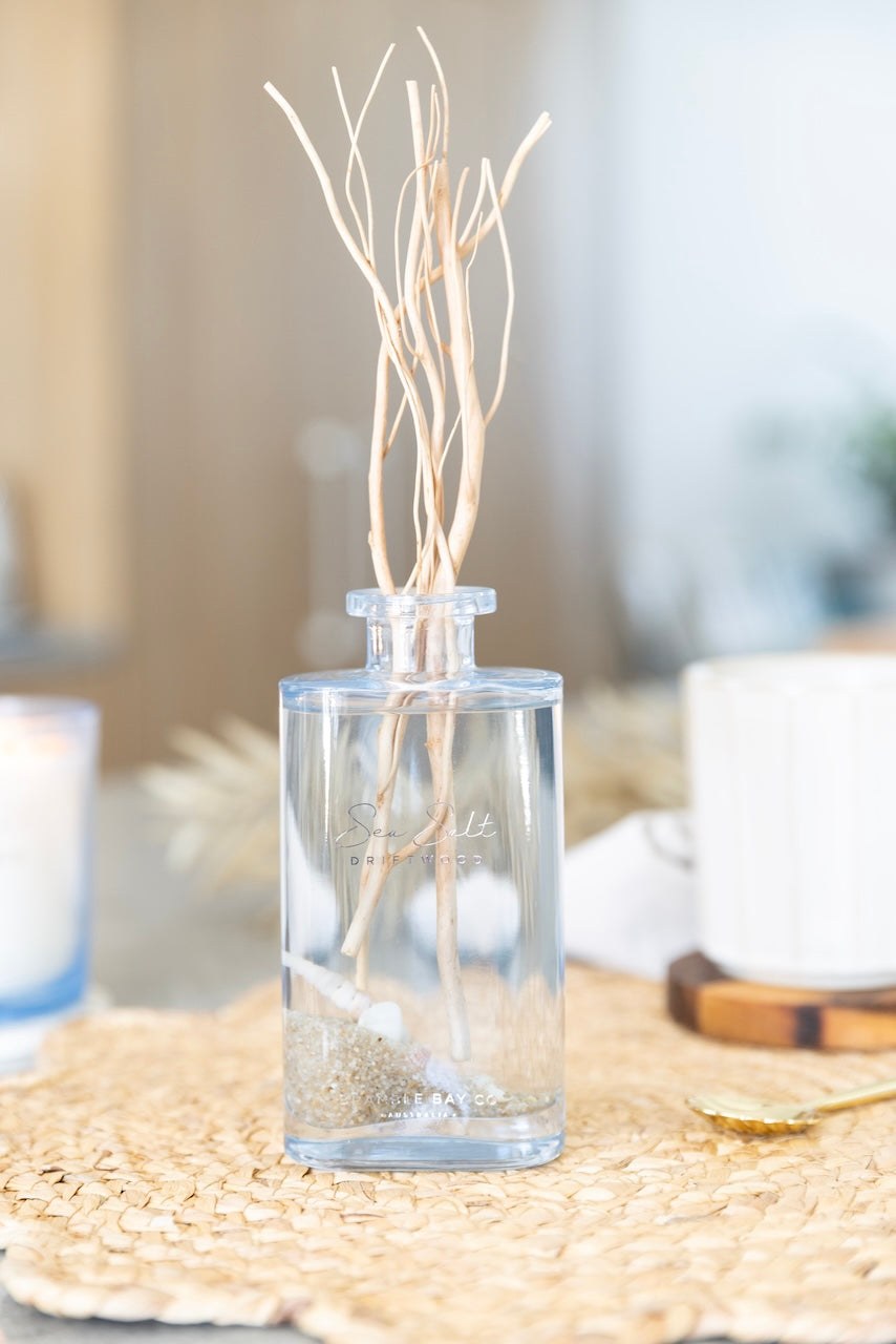 The glass diffuser bottle with willow reeds, sand and shells, standing on a woven mat.