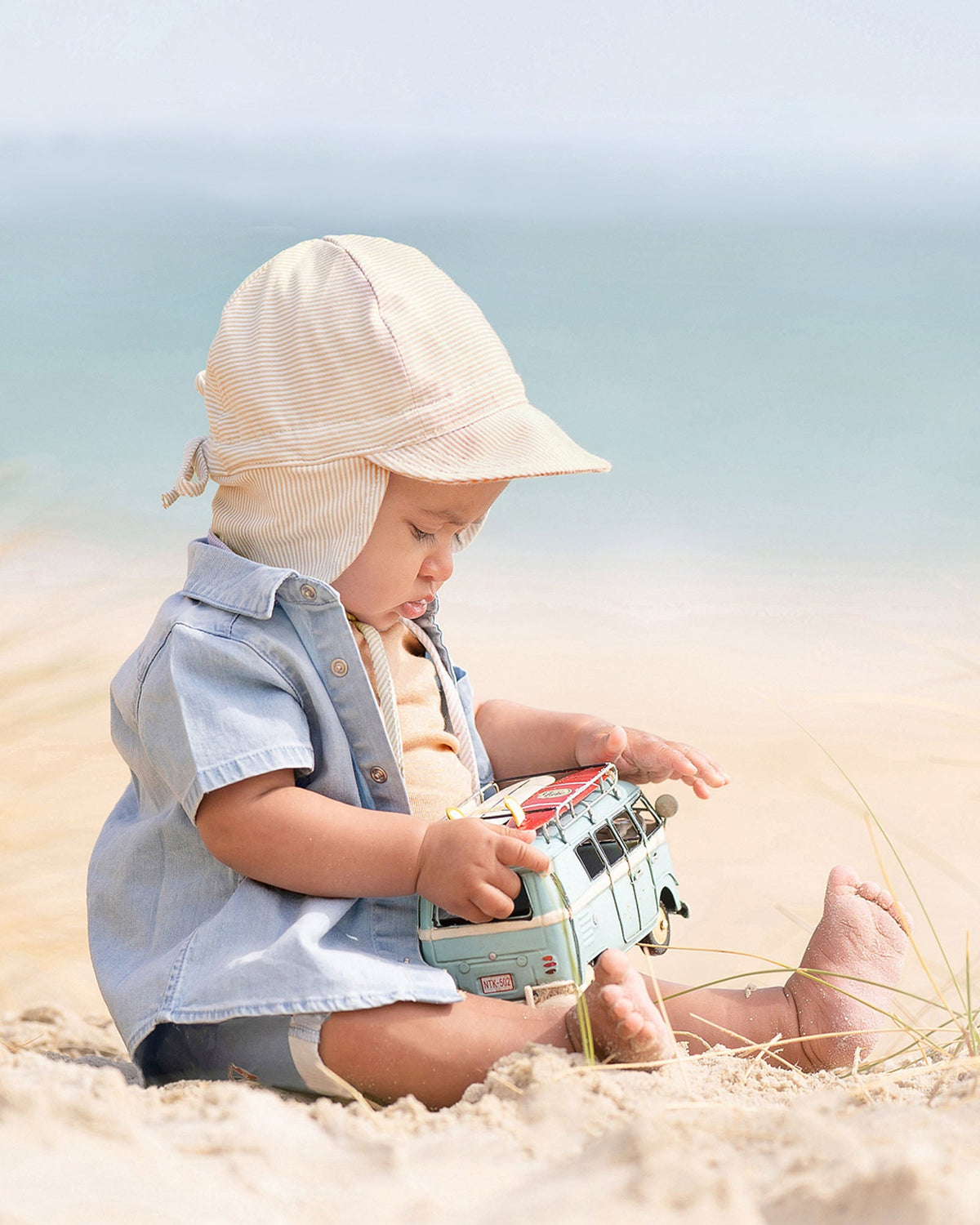 Baby on the sand in the Indiana shirt and a cream flap cap, playing with a toy van