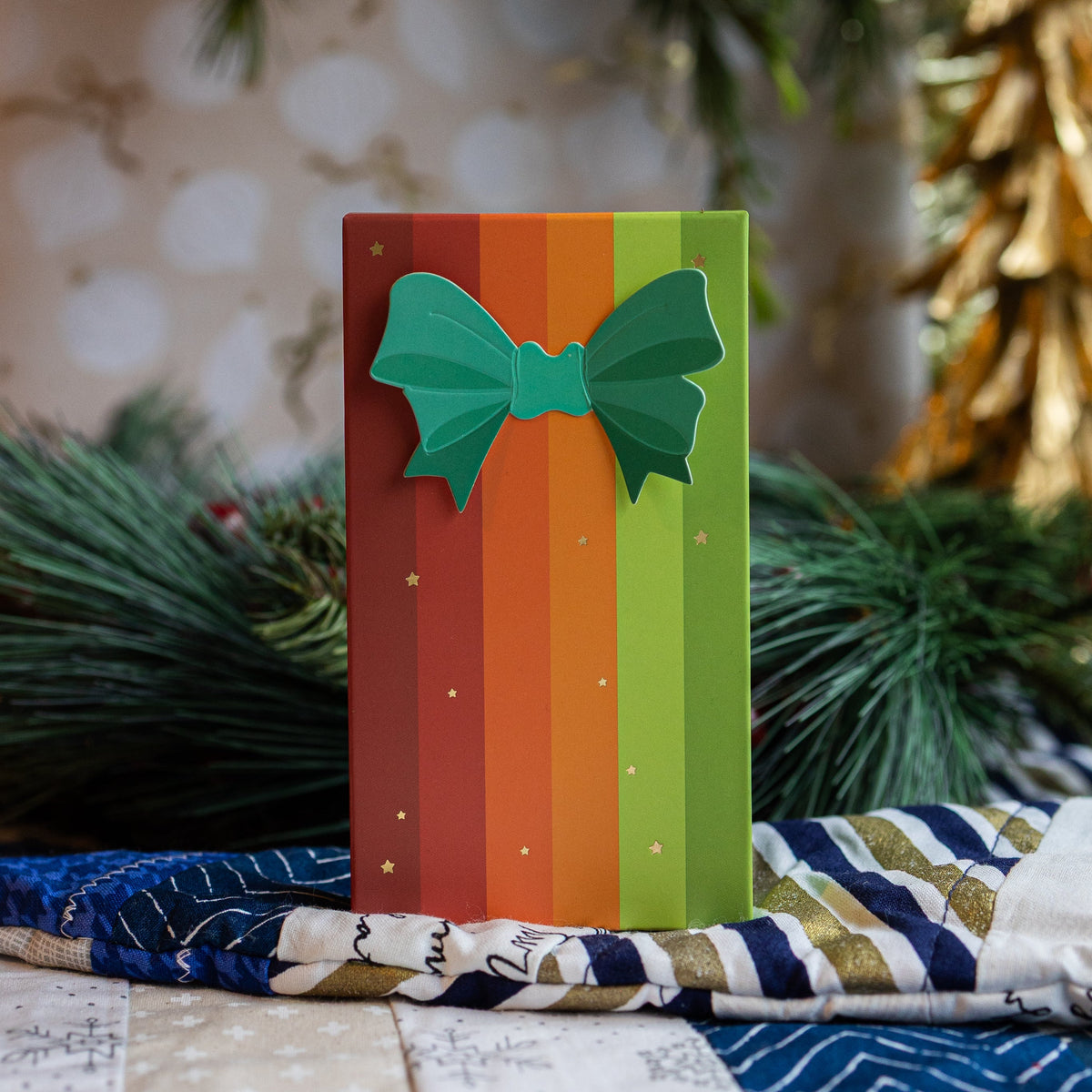 Warm and Bright gift box in red, orange and green stripes with a green bow, standing in front of pine branches.