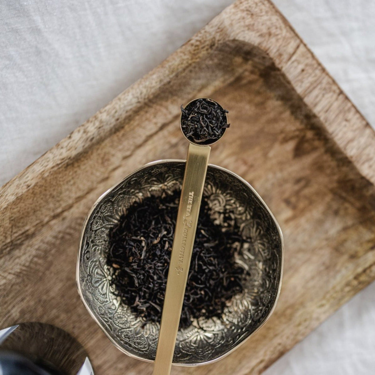 Gold Tea Collective spoon holding black tea leaves over an engraved bowl of loose leaf on a timber board