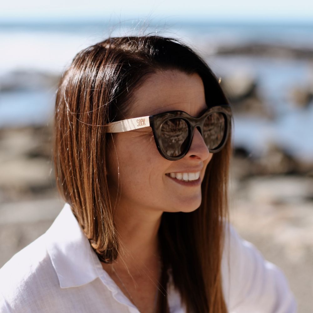 Close view of a woman with long brown hair smiling in the Milla Khaki sunglasses, rocks and sea behind her