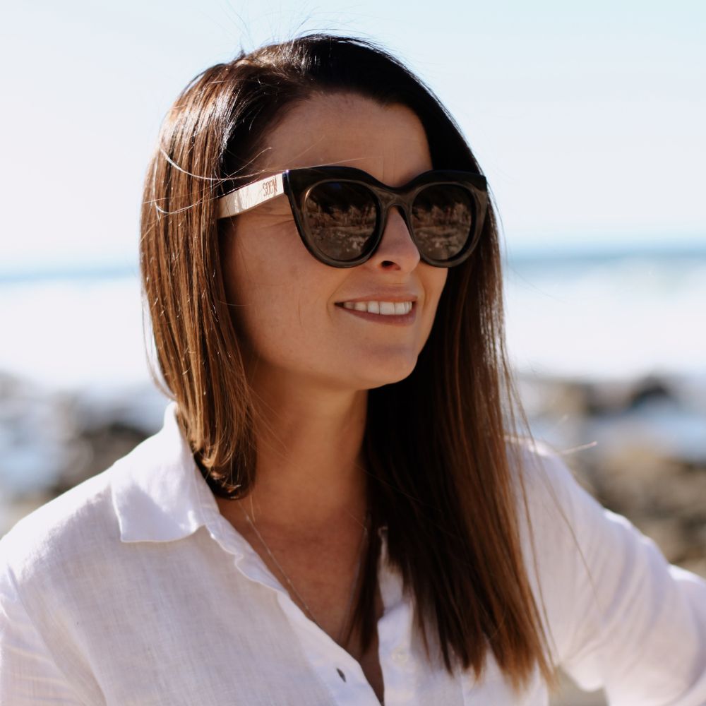 A woman with long brown hair looking off to the side in the Milla Khaki sunglasses, the sea behind her