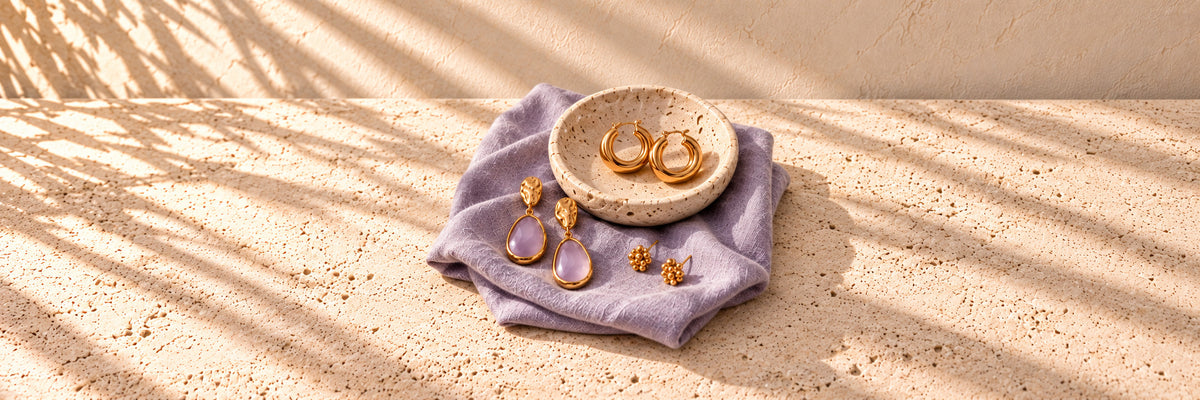 Gold hoops in a stone dish and lilac drop earrings on folded lavender linen