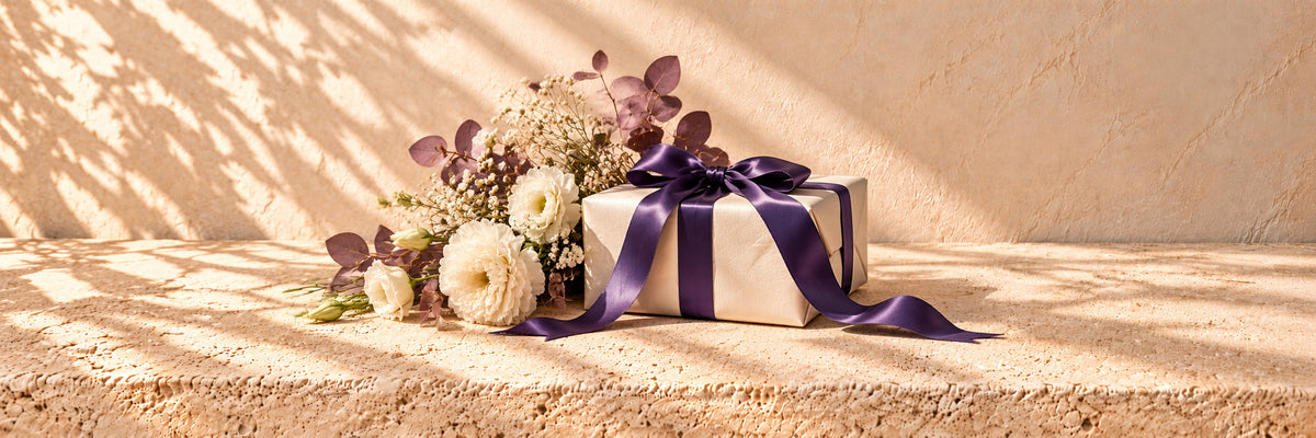 An ivory parcel with a purple satin bow beside ivory flowers and mauve foliage