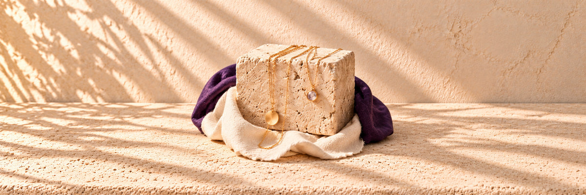 Fine gold chains with small pendants draped over a limestone block on ivory and purple cloth
