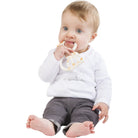 Baby in a white top and grey leggings sitting and chewing the ring teether.