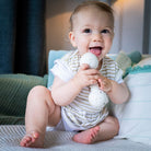 Baby sitting on a blanket, holding the cream rattle in both hands and mouthing the lower ball.