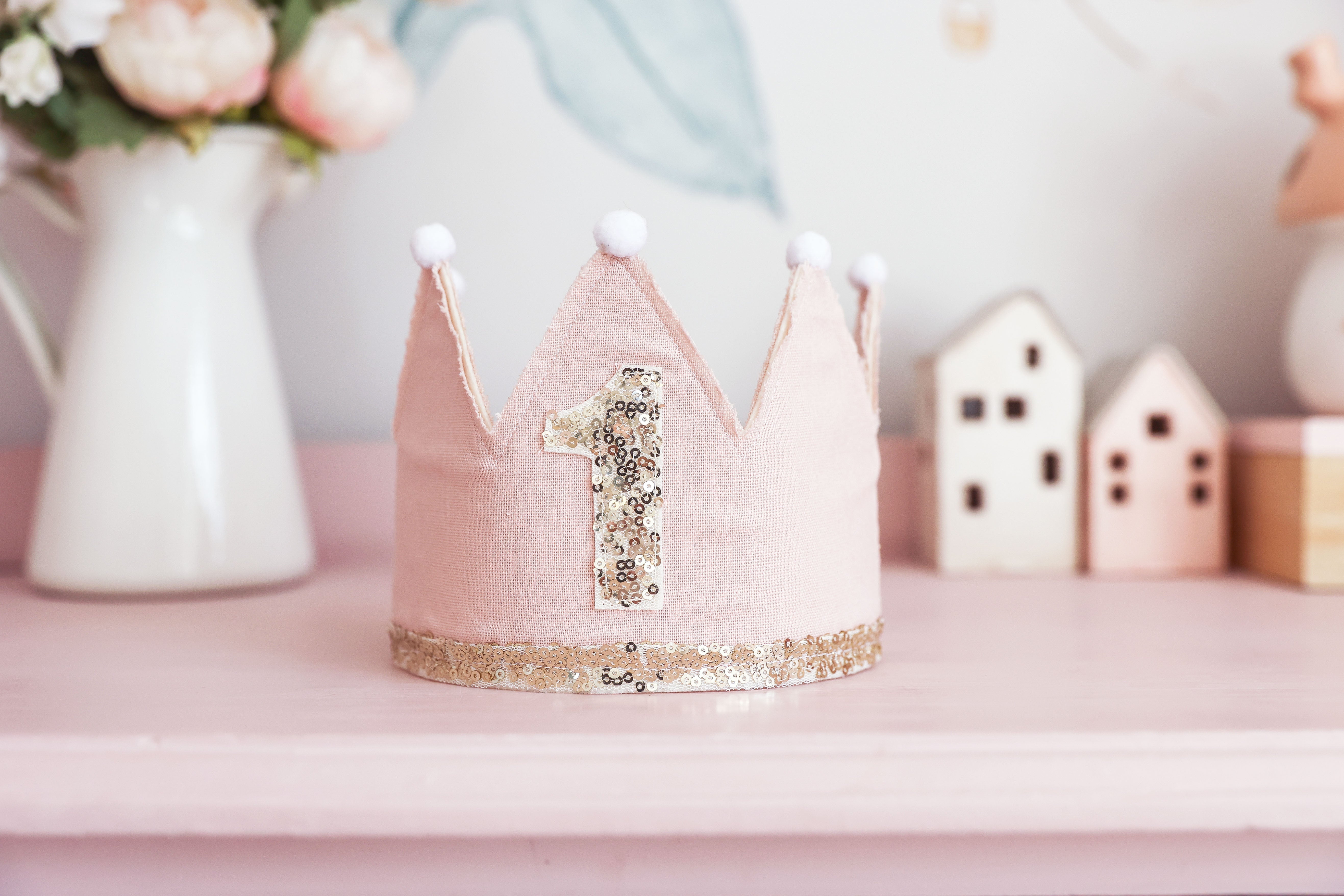 Pink first birthday crown on a shelf with flowers and little wooden houses
