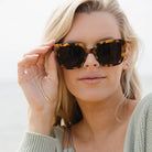 A woman with long blonde hair lifting the Soluna sunglasses, worn outdoors