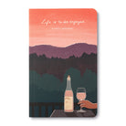 Journal cover of a coral sunset over layered hills with a hand raising a glass of wine