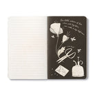 Open journal with lined pages beside a black page of white poppies, scissors, an envelope and an inkpot, with a line about little actions of love adding up