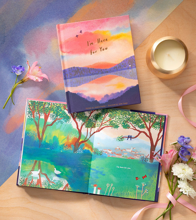 The book styled on a table beside a candle and small purple flowers, opened to a green valley spread