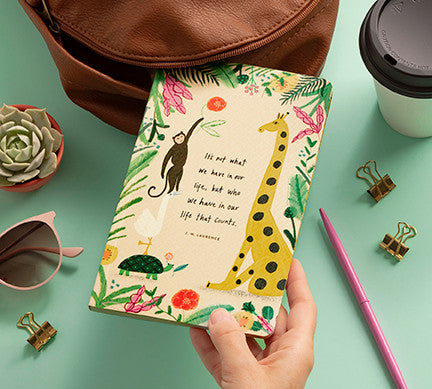 Hand holding the jungle-animal journal on a desk beside a satchel, succulent and pink pen
