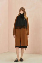 Full-length camel coat look with the Ebony black scarf, pink wall behind