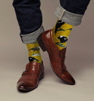Man's feet in brown leather shoes with turned-up jeans, wearing the mustard magpie socks against a grey backdrop.