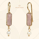 Euro Gold earrings with pale pink strawberry quartz bars and freshwater pearl drops, on patterned cream backdrop
