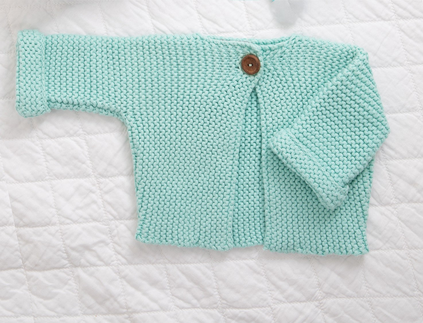 Aqua cardigan laid flat on a white quilted blanket, one sleeve folded across.