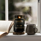 Lit Campfire candle on a wooden coaster at a rainy windowsill beside an open book and a stoneware mug