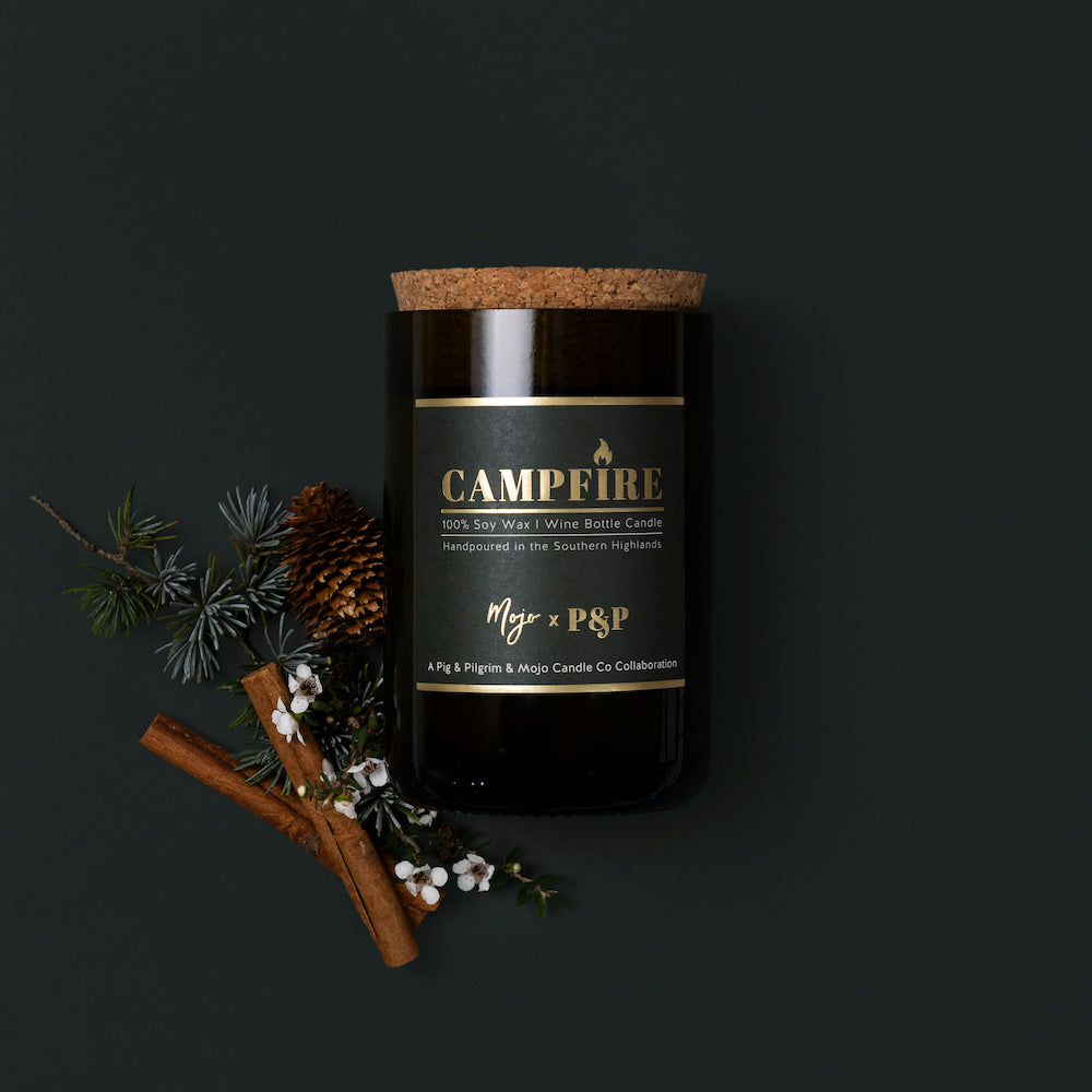Campfire candle in a dark reclaimed bottle on charcoal, with pine sprig, pinecones and cinnamon sticks