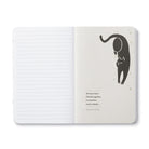 Open journal with lined pages beside a cream page of a stretching black cat above a short quote