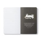 Open journal with lined pages beside a charcoal breakout page of a white leaping cat and a quote