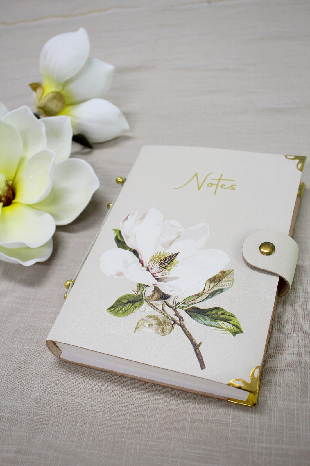 Magnolia leather notebook on a linen cloth beside two white magnolia flowers