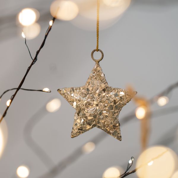 Star ornament covered in chunky champagne flakes, hanging from a ring on a gold cord among warm fairy lights