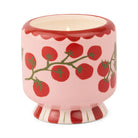Pink ceramic candle painted with red tomatoes on green vines, a red scalloped rim and a red and white striped foot, lit.