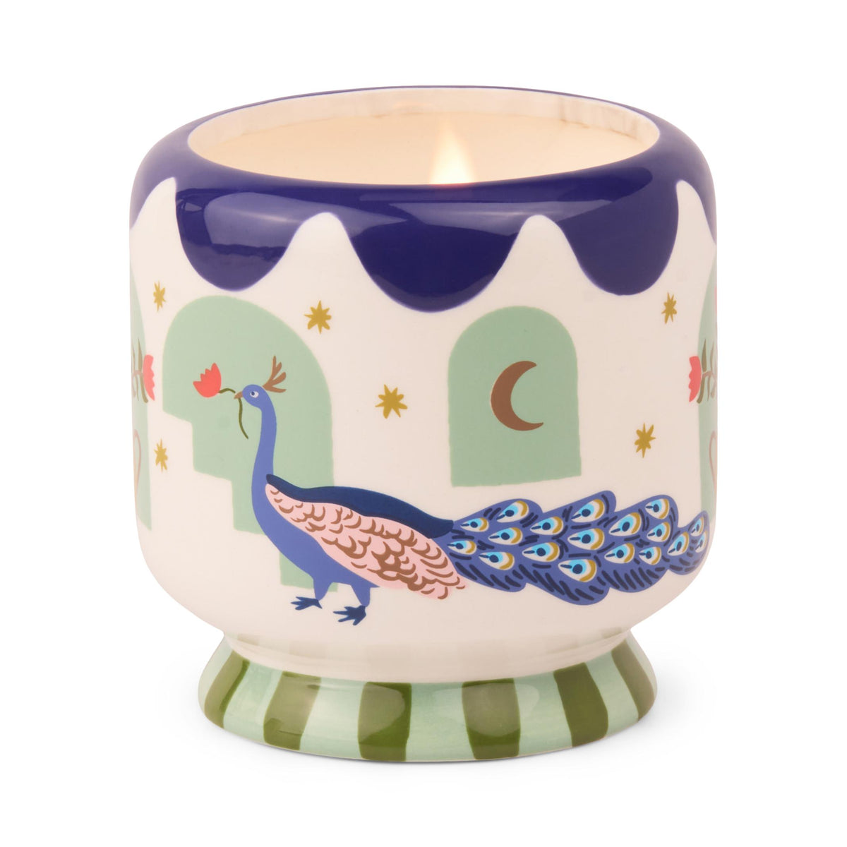 Small cream ceramic candle with a navy scalloped rim and a green striped foot, painted with a peacock carrying a red flower past mint arches, lit.