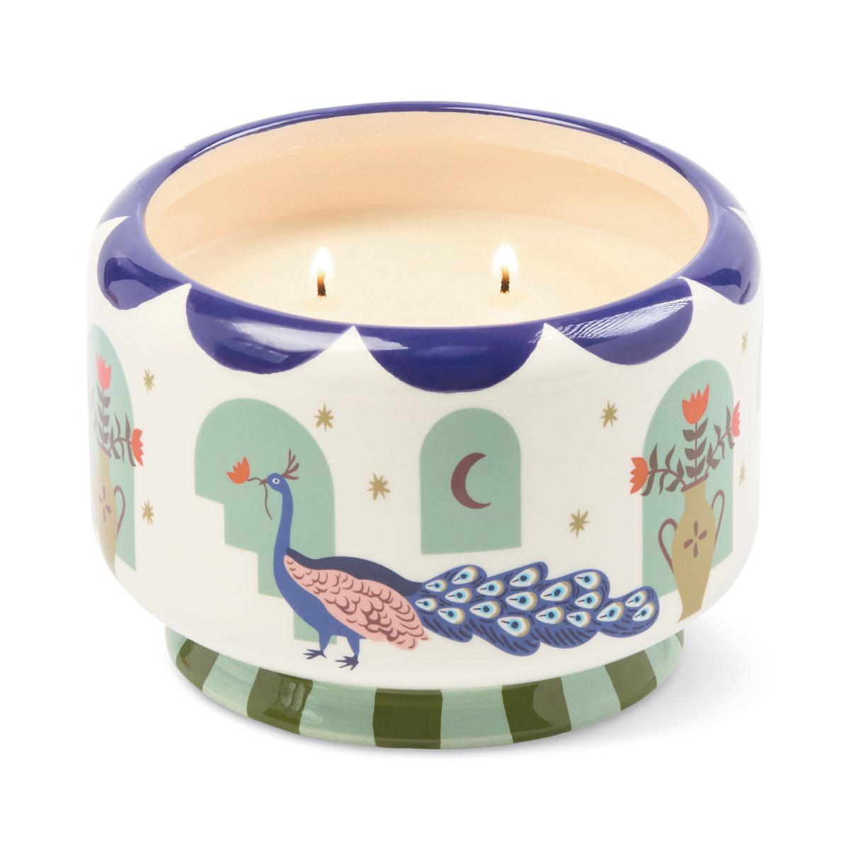Wide cream ceramic candle with a navy scalloped rim, painted with a peacock and mint arches, its two wicks lit.