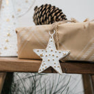White metal star ornament embossed with small gold stars and dots, hung by a jute loop on a kraft-wrapped gift with a pine cone behind