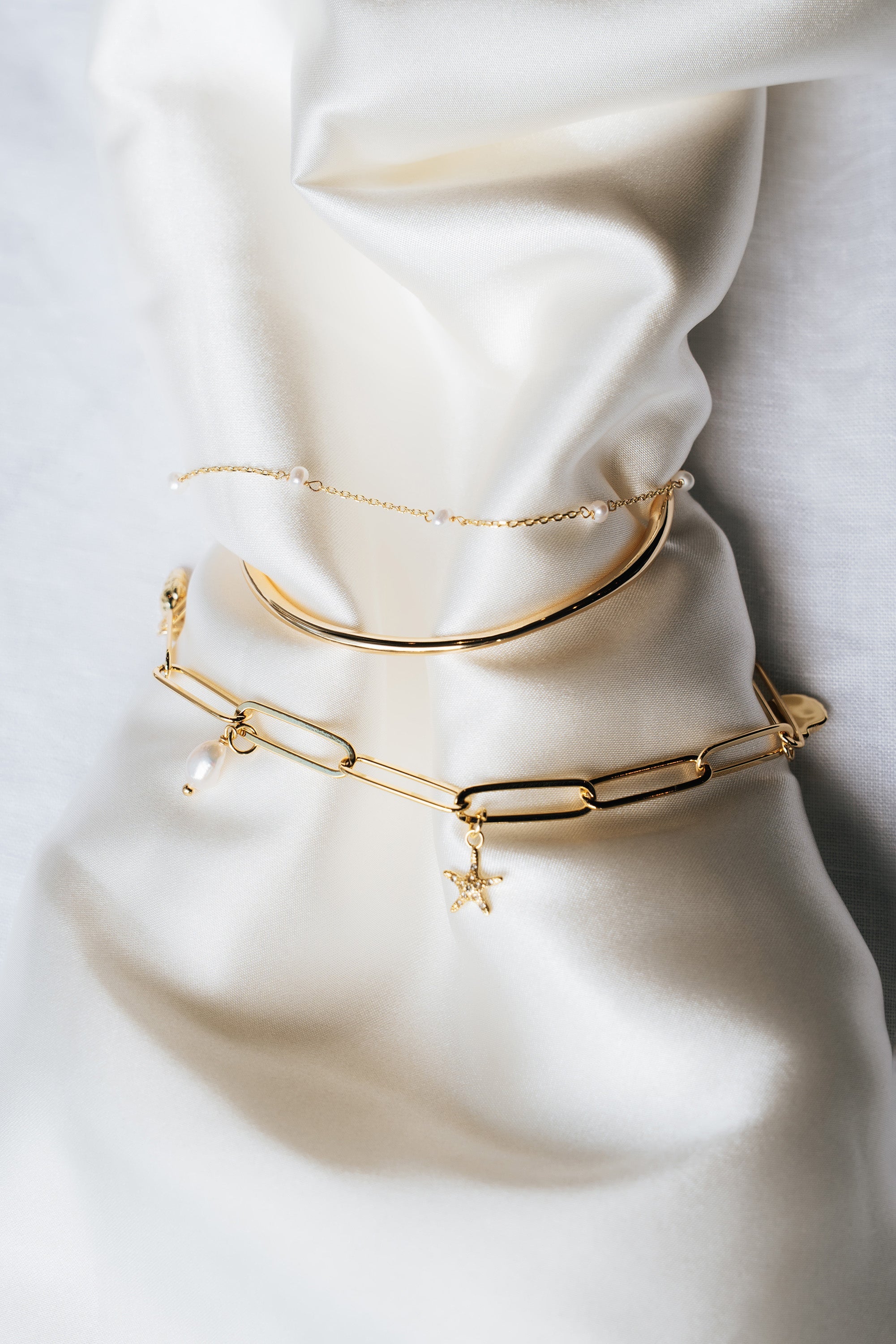 The soft gold Louella bangle layered with fine gold chain bracelets on cream silk.