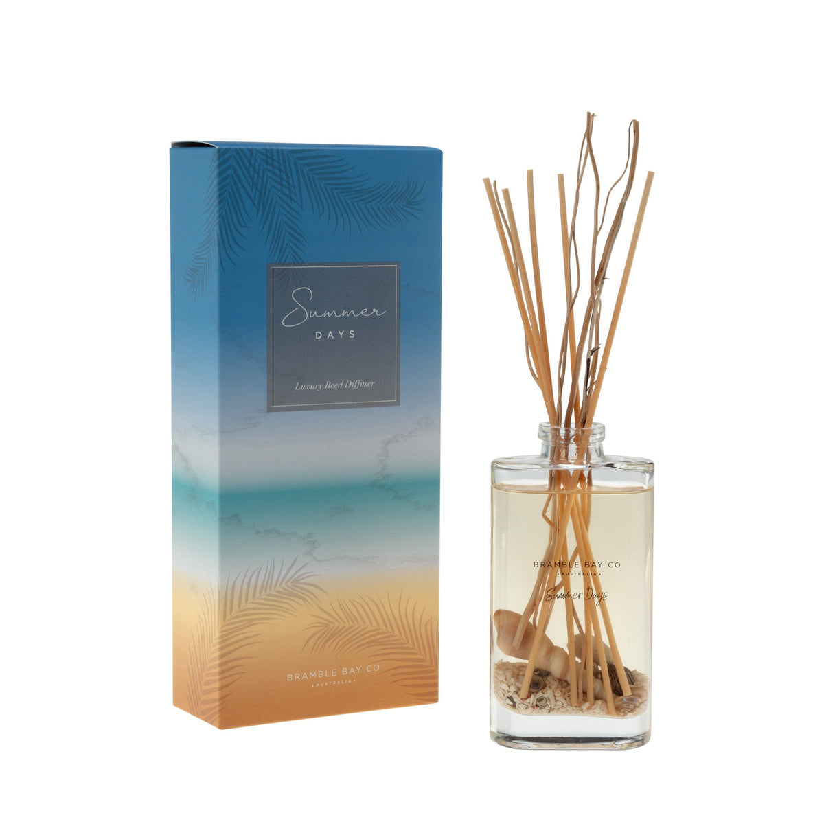 Summer Days gift box in blue-to-orange beach ombre with palm print, beside a glass reed diffuser with willow reeds, sand and shells