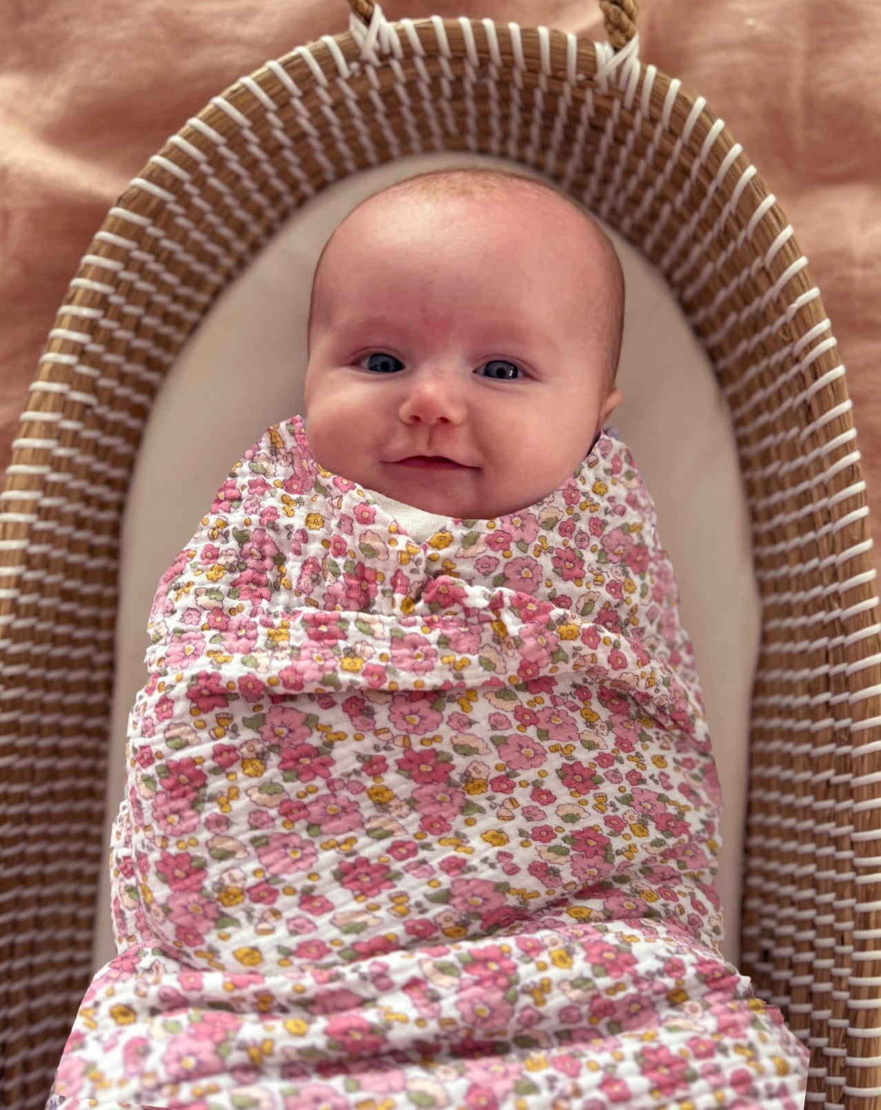 Baby swaddled in rose floral muslin lying in a woven bassinet